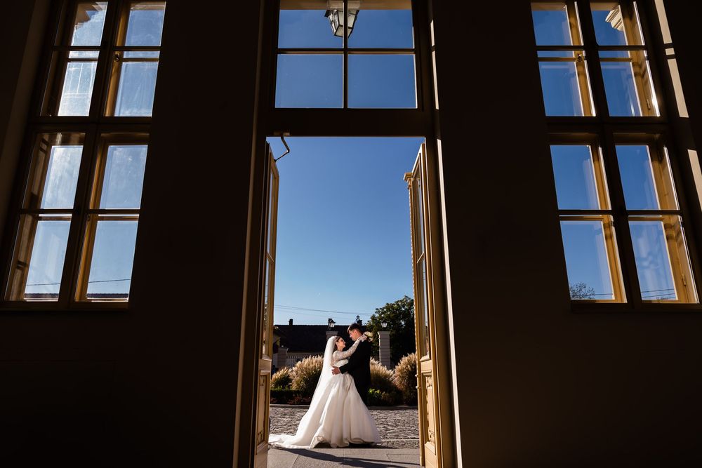 Bride and groom silhouetted in elegant doorway, sharing romantic moment under blue sky at outdoor wedding venue.
