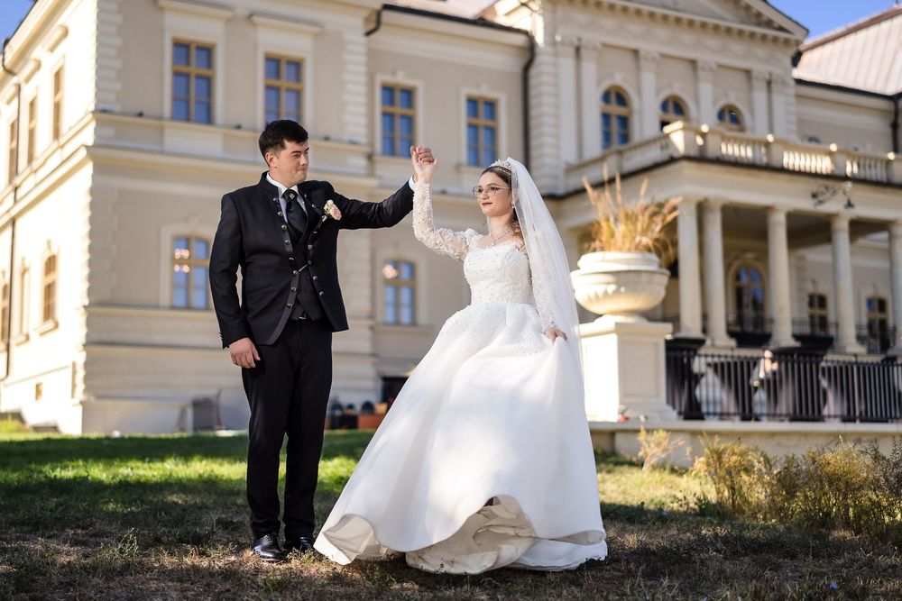 Bride and groom dancing outdoors in front of elegant manor, bride in white gown with veil, groom in black suit.