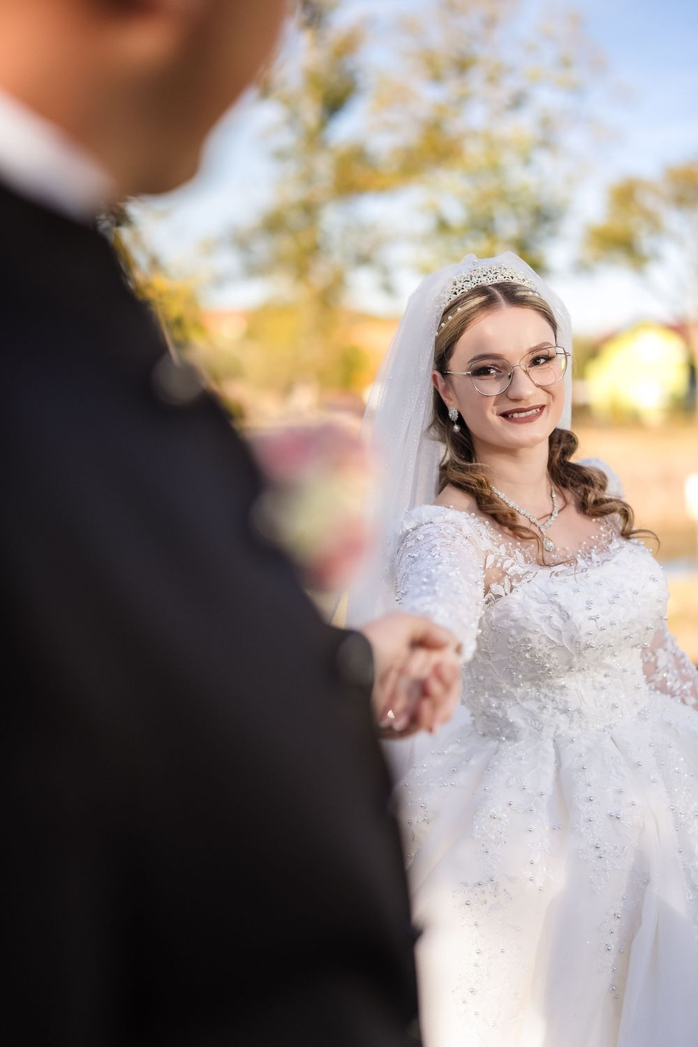 Smiling bride in white gown and tiara holds groom's hand outdoors on sunny wedding day.