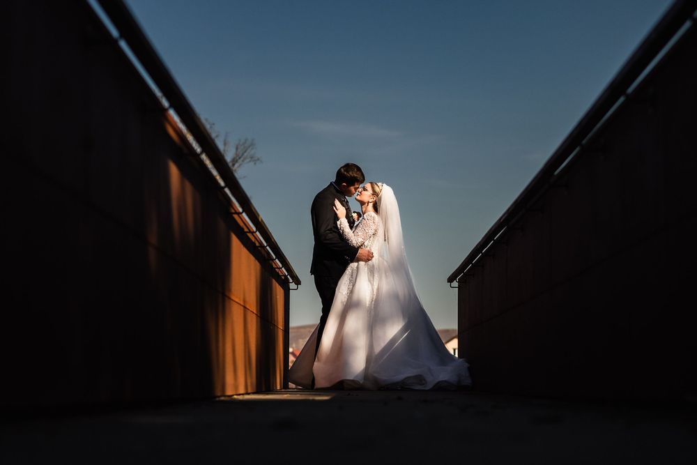 Bride and groom share a romantic kiss at golden hour, framed by dark walls against a blue sky.