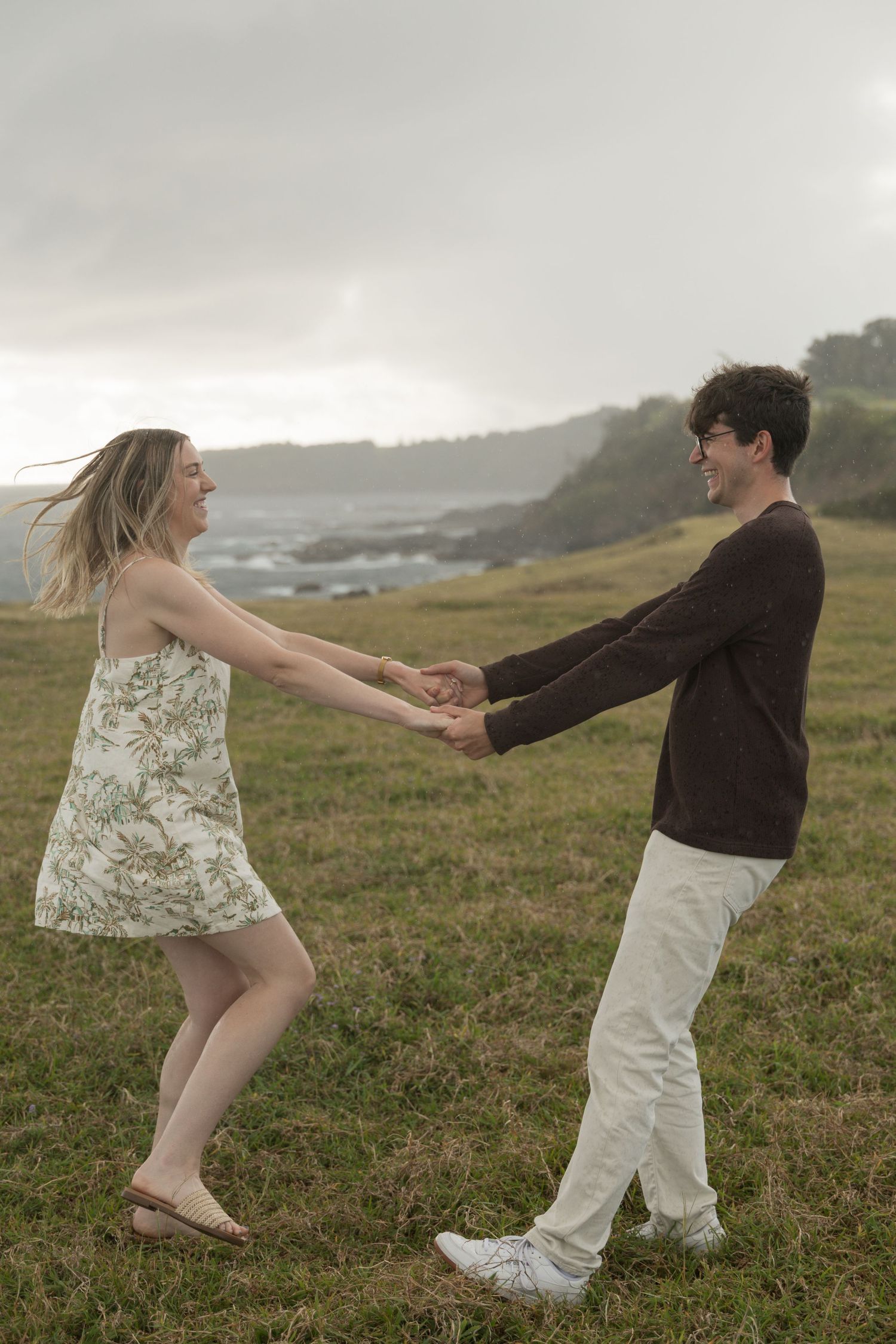A couple holds hands and dances joyfully in a coastal field, with ocean cliffs visible in the misty background.