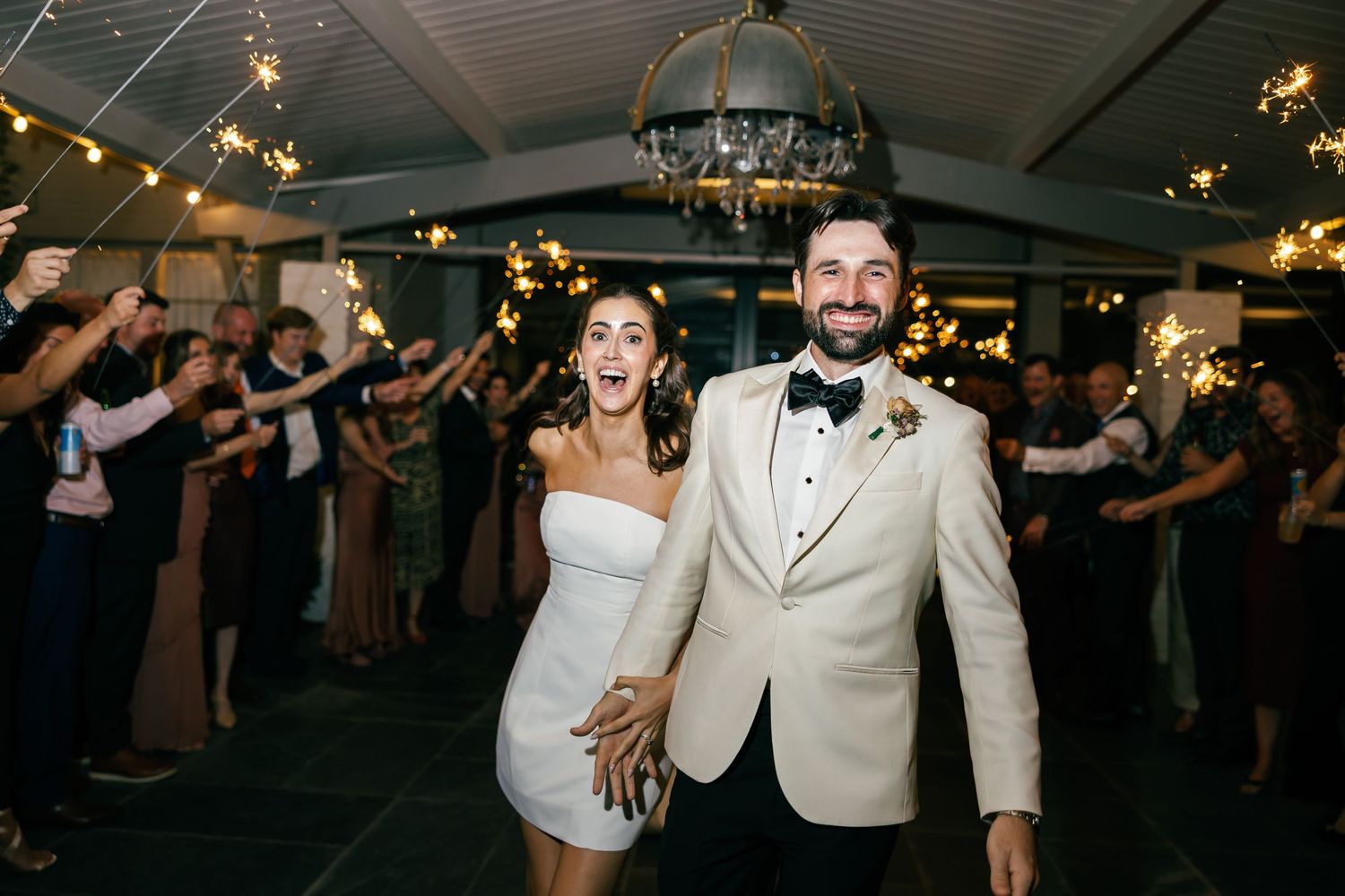 Bride and groom exit wedding reception through sparkler tunnel, smiling joyfully, groom in white tuxedo jacket.