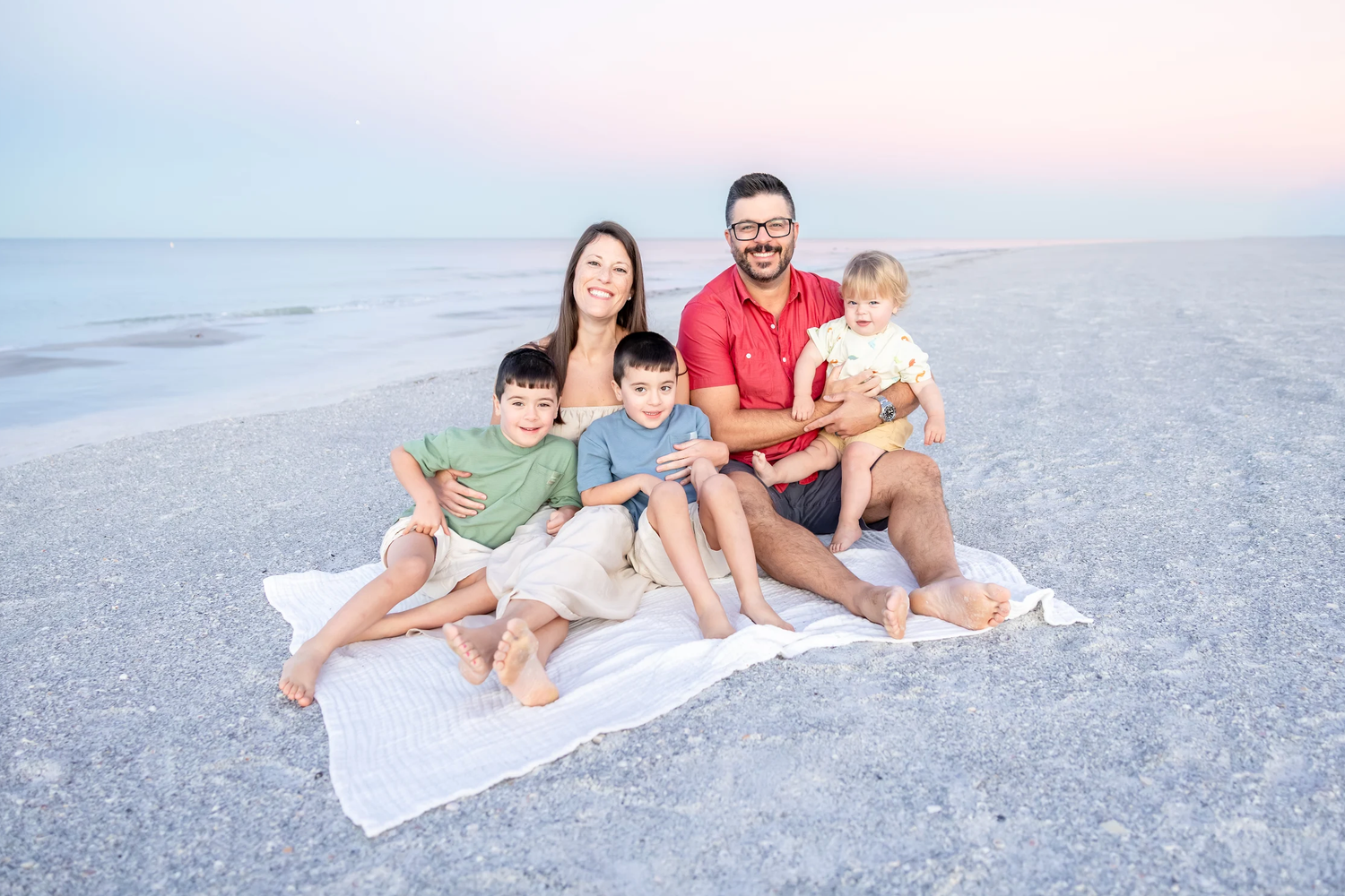 Family of five smiling together on a white blanket at the beach during sunset.