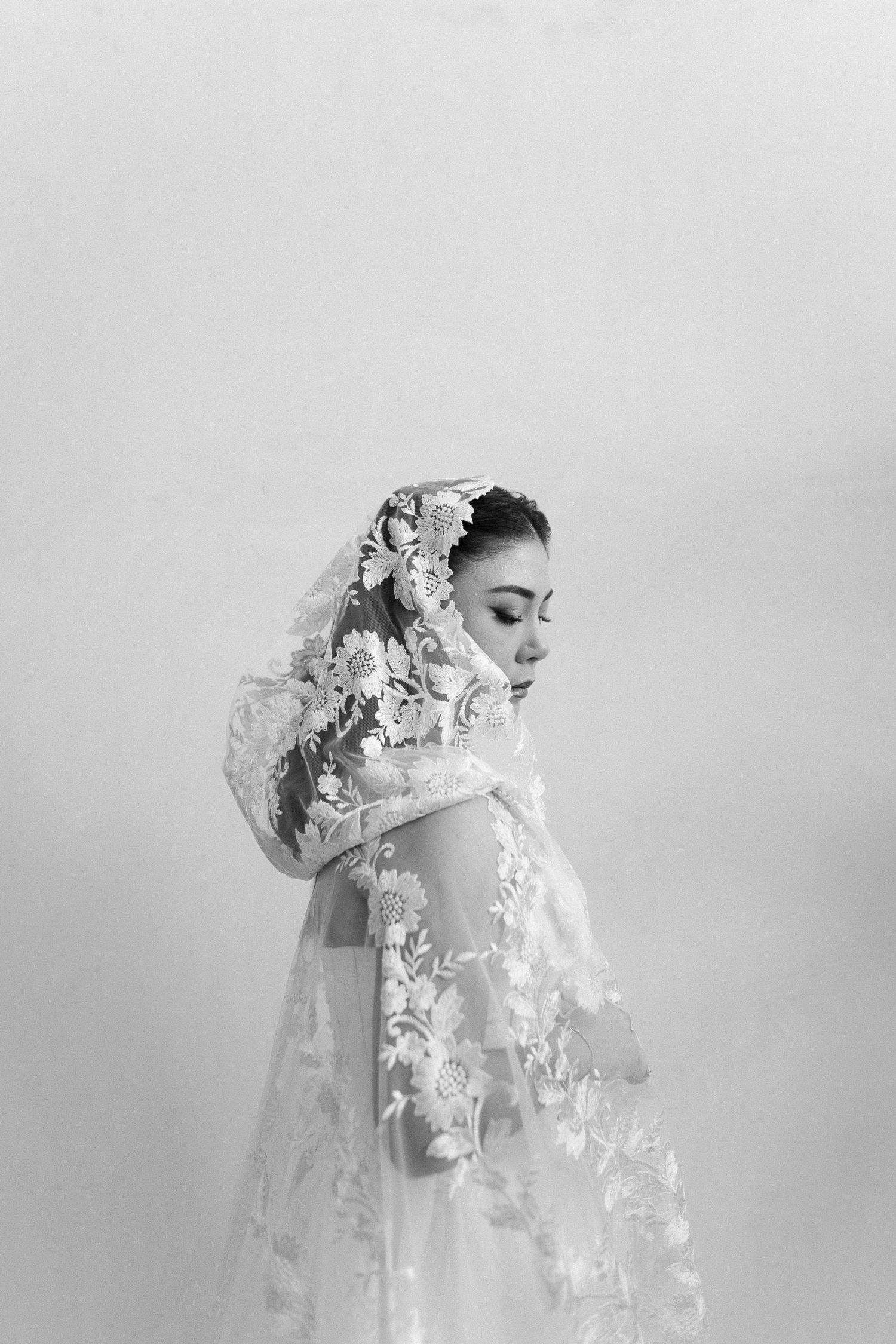 Elegant bride in a white lace gown and floral veil, posing gracefully in a black and white portrait.