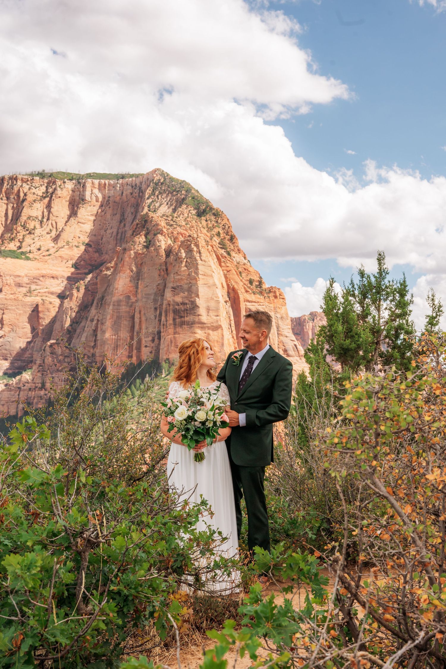 A couple in wedding attire holds hands outdoors at Zion National Park, with red rock cliffs and pine trees behind them.