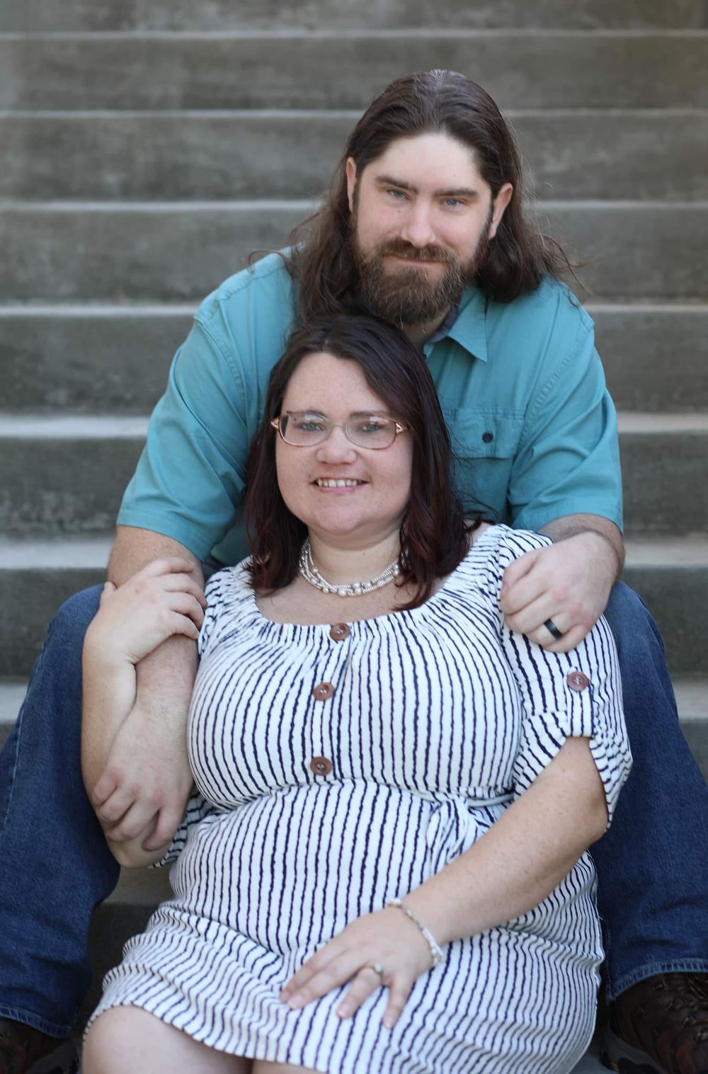 Happy couple posing on outdoor stone steps, woman in striped dress, man with long hair.