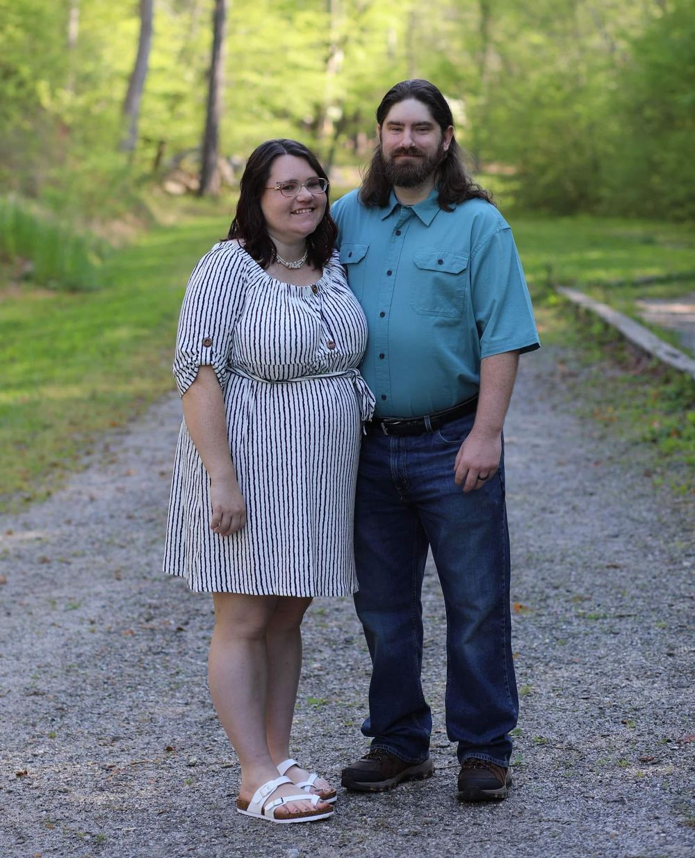 Smiling couple standing together on a gravel path surrounded by lush green trees.