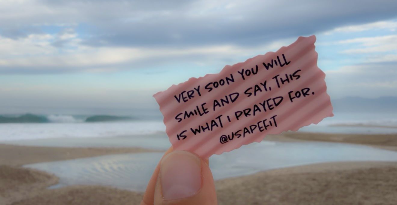 Hand holding a pink note with inspirational quote on a beach, ocean waves and cloudy sky in background.