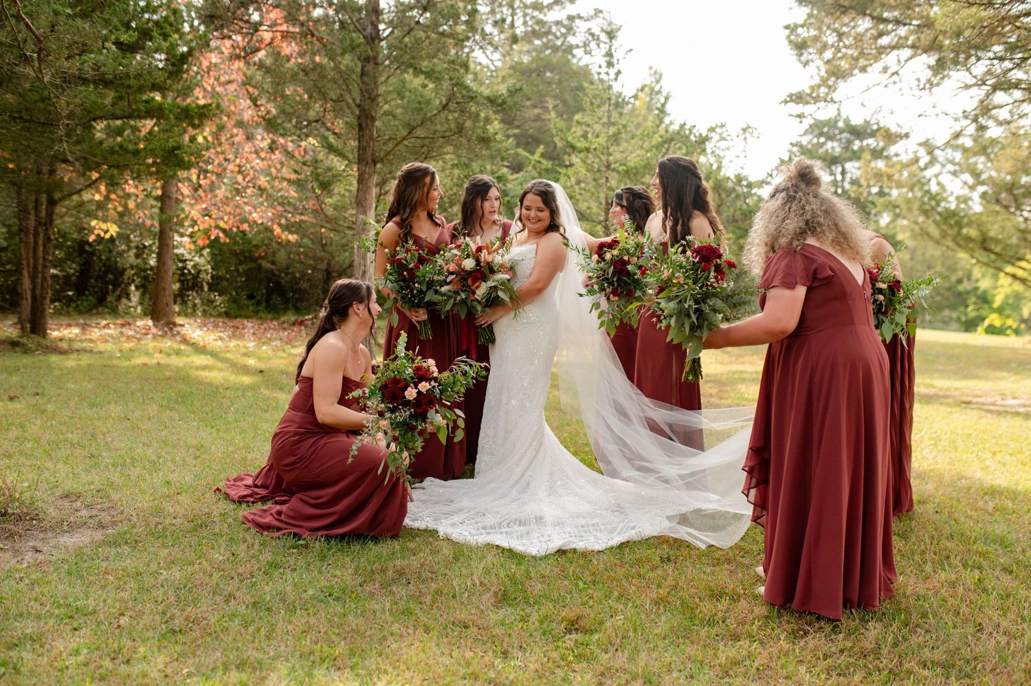 Bride with bridesmaids in burgundy dresses admiring bouquets of red and green flowers on a lawn.