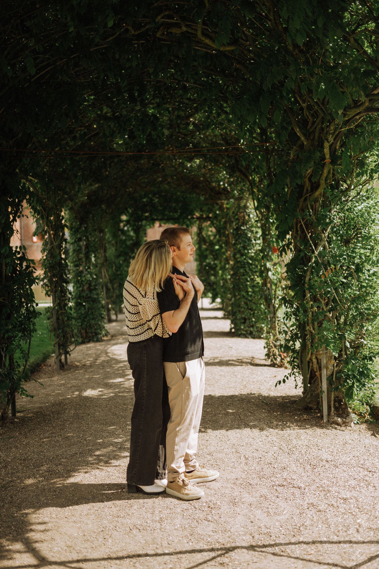 Couple embracing and gazing into the distance beneath a shaded green garden pergola with dappled sunlight on a gravel path. De Haar