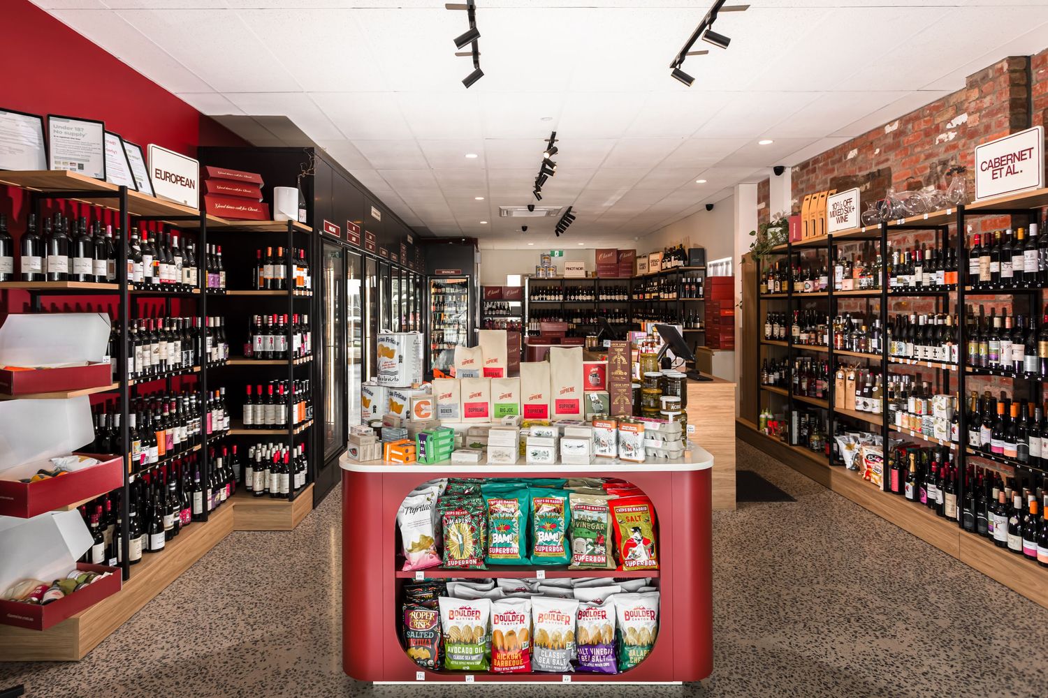 Interior of a modern bottle shop with wine racks lining the walls and a red central display counter stocked with packaged snacks.