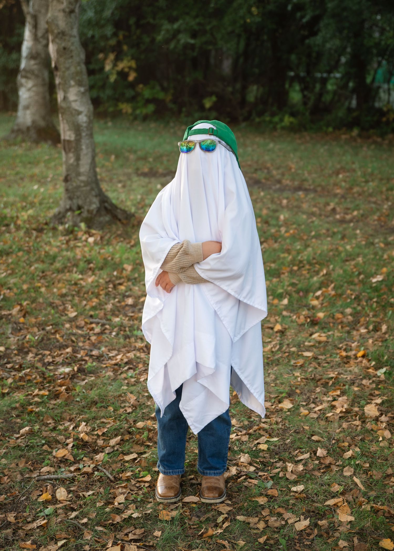 Child in a white sheet ghost costume with green hat and sunglasses stands on a leaf-covered lawn near trees in autumn.