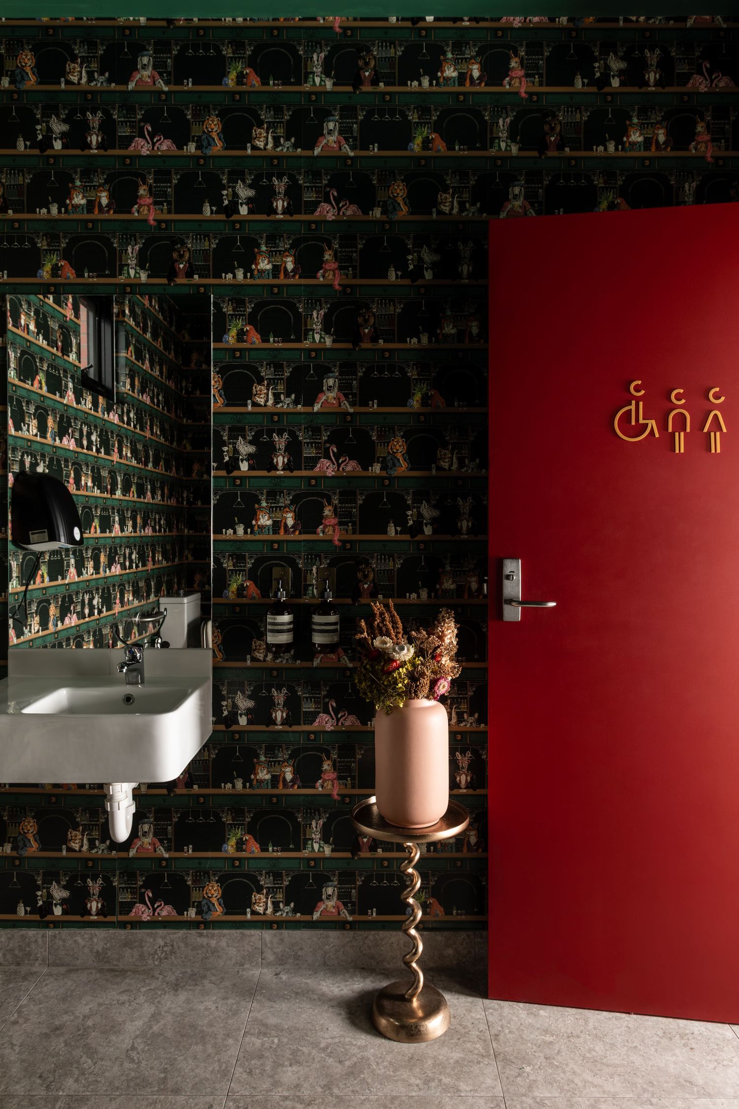 Stylish restroom with dark patterned wallpaper, white wall-mounted sink, red door with gold accessible signage and a pink vase on a twisted 