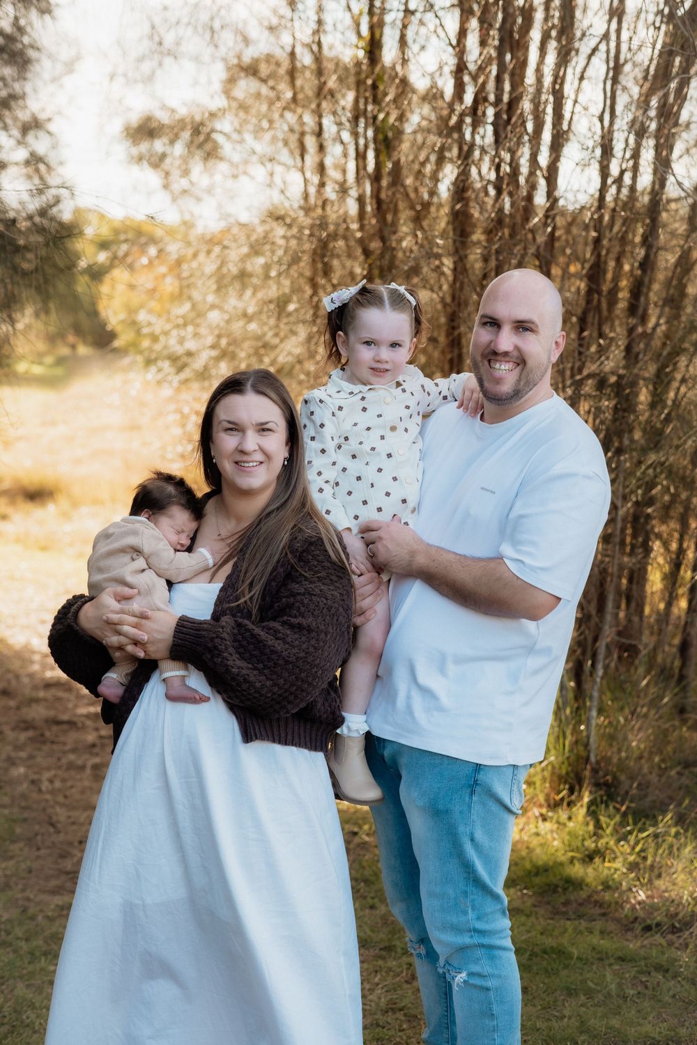 Smiling couple holding their baby and toddler in a sunlit outdoor field with tall trees, posing for a warm family portrait in Rouse Hill.