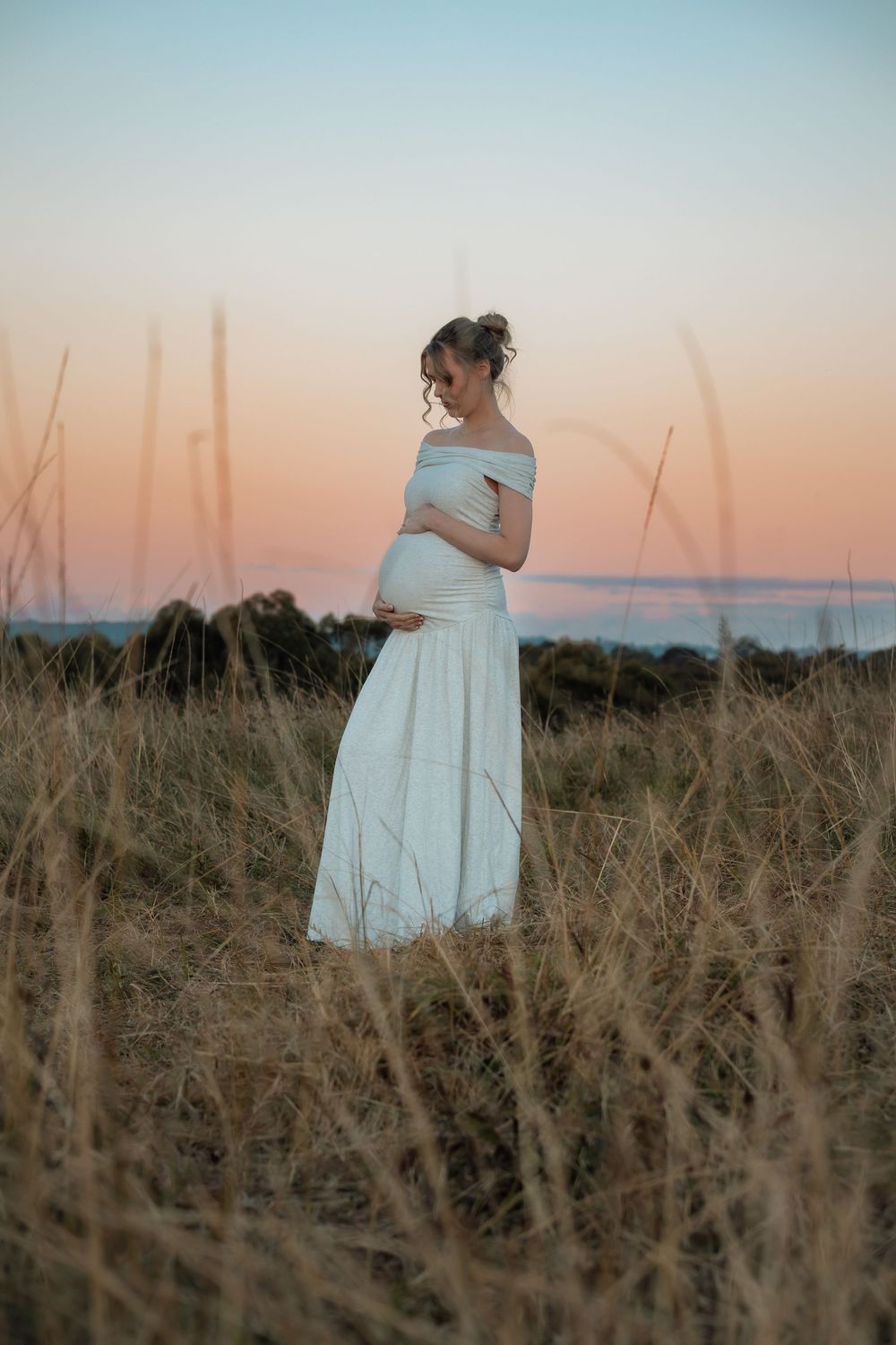 Pregnant woman in a white off-the-shoulder maternity gown cradling her belly in a tall grass field at sunset in Rouse Hill Regional Park.
