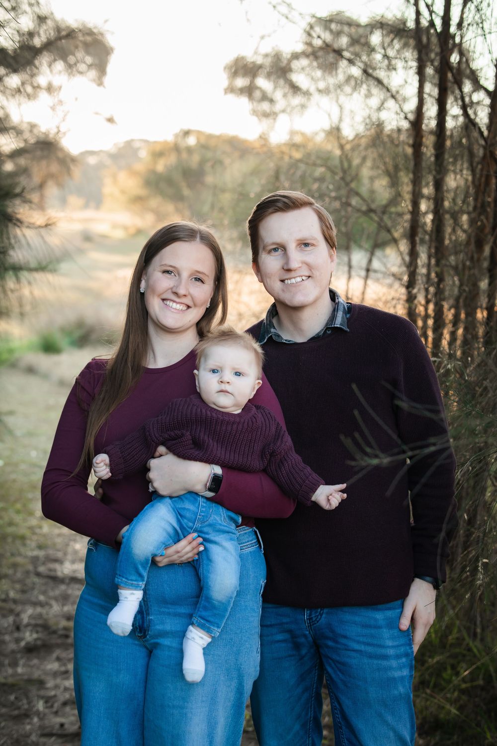 Smiling couple in burgundy sweaters and jeans holding their baby outdoors in a sunlit bushland setting for a family portrait in Rouse Hill.