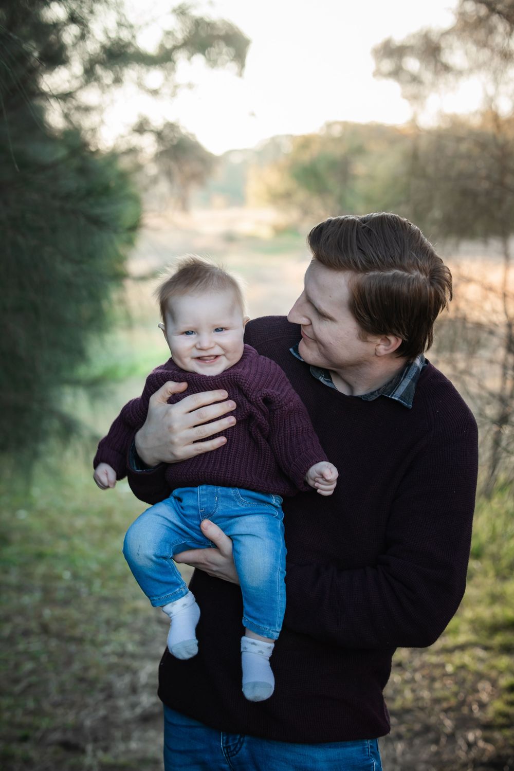 Smiling baby in a maroon sweater and jeans is held up by a man in a dark sweater on a grassy path with blurred trees behind in Rouse Hill.
