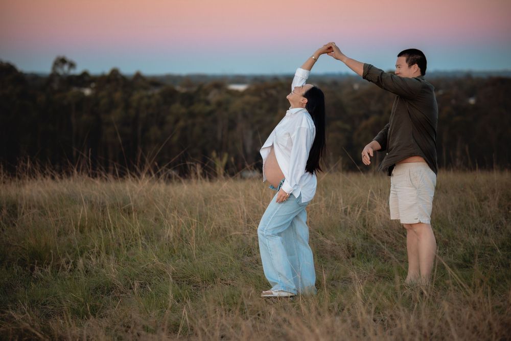 Pregnant woman in white shirt and jeans twirls under her partner's raised arm while dancing in a grassy field in Rouse Hill at sunset.