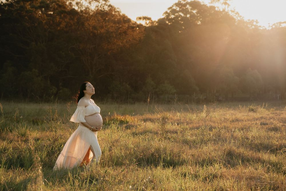 Pregnant woman in a flowing cream dress cradles her belly in a golden meadow at sunset for a maternity photoshoot in Rouse Hill.