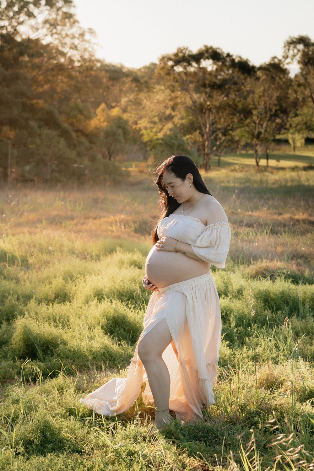 Pregnant woman in a flowing cream off-shoulder gown holding her belly in a sunlit grassy field in Rouse Hill, a golden hour maternity photo.