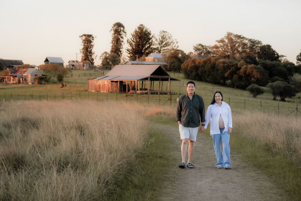 Couple holding hands and walking on a dirt path through golden grass toward a rustic wooden barn at sunset in the countryside in Rouse Hill.