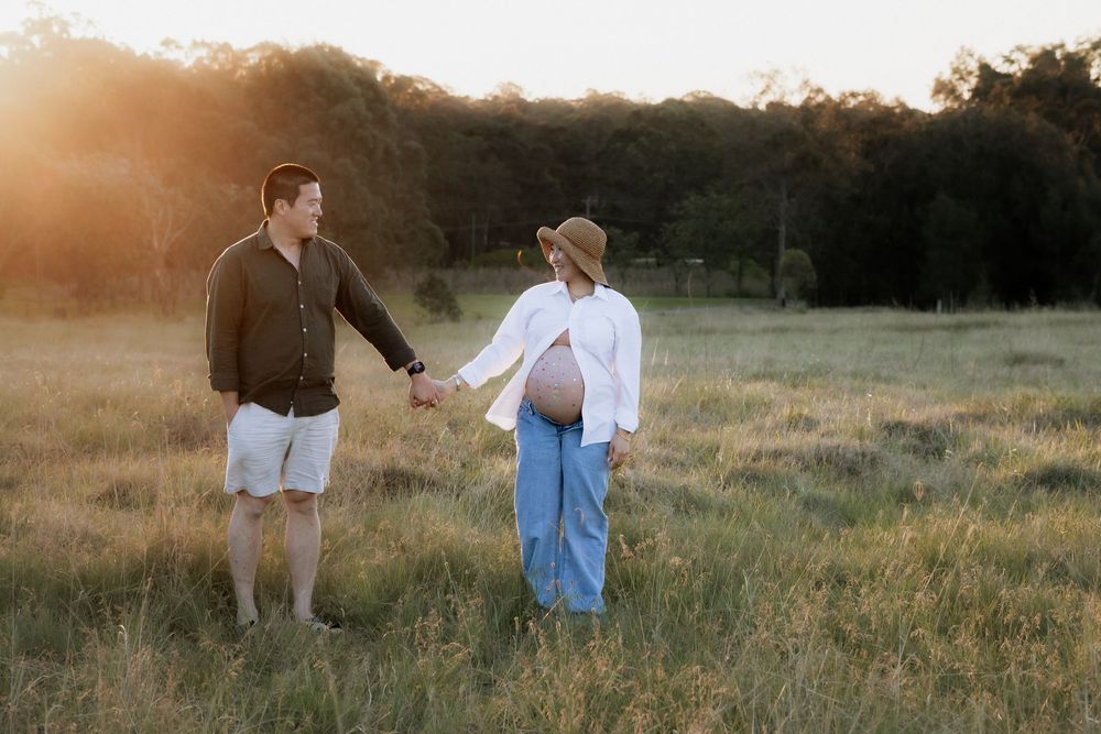 Expectant couple holding hands in a golden grassy field at sunset, the pregnant woman in a straw hat showing her baby bump in Rouse Hill.