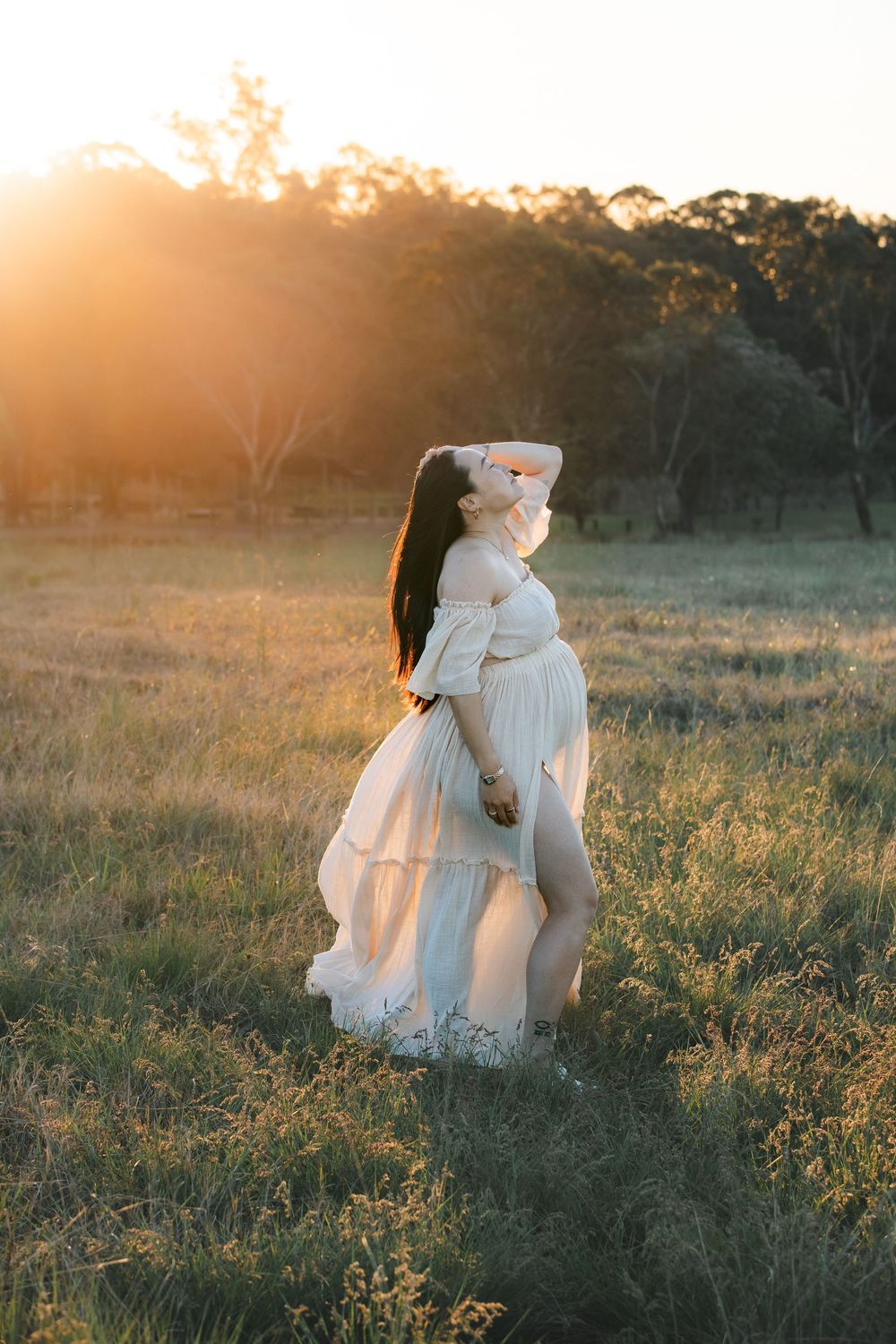 Pregnant woman in a flowing white off-the-shoulder dress poses in a grassy field at golden hour for a sunset maternity photoshoot.