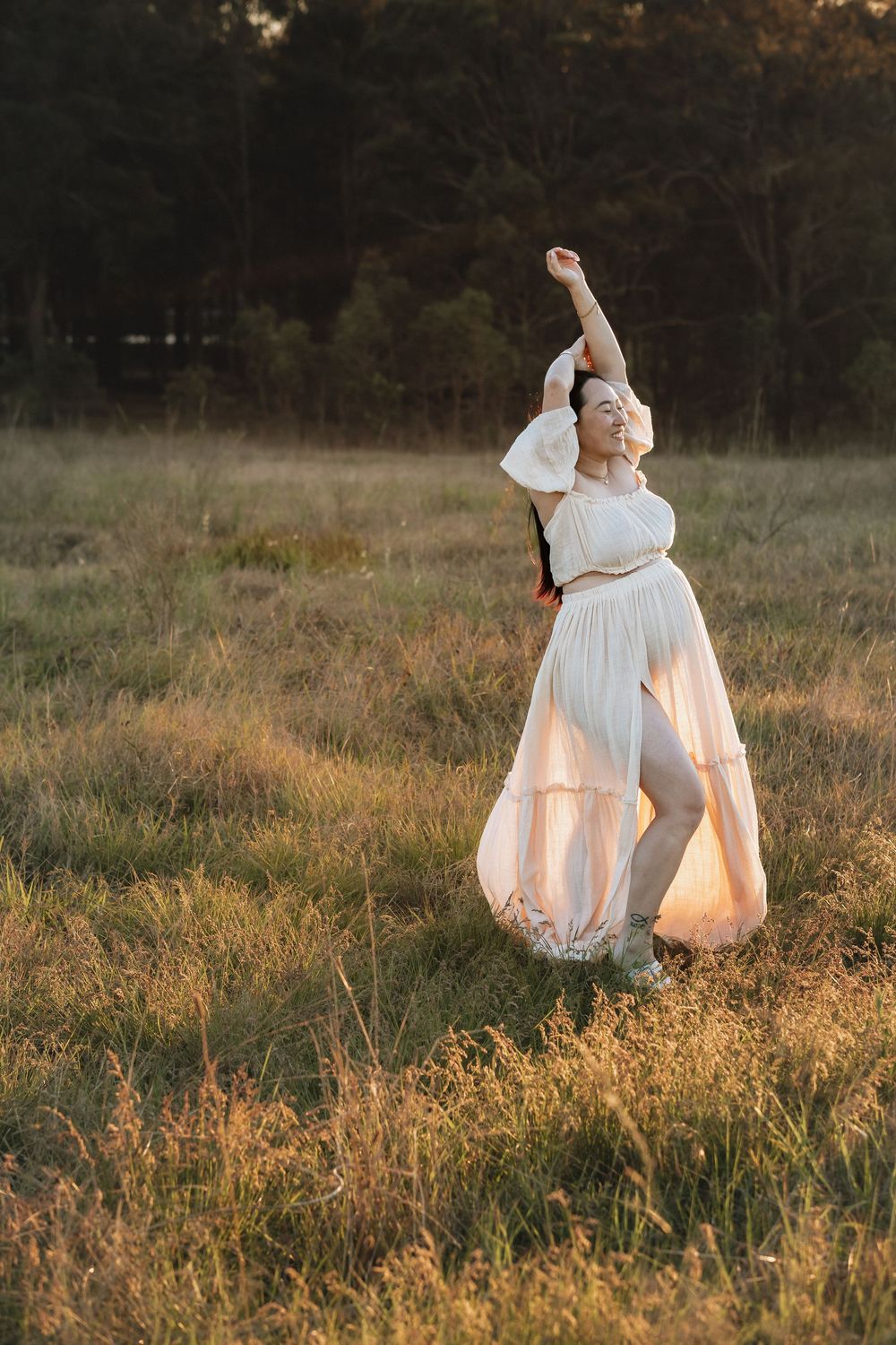 Woman in a flowing white dress raises one arm and smiles while standing in a golden grassy field at sunset with trees in the background.