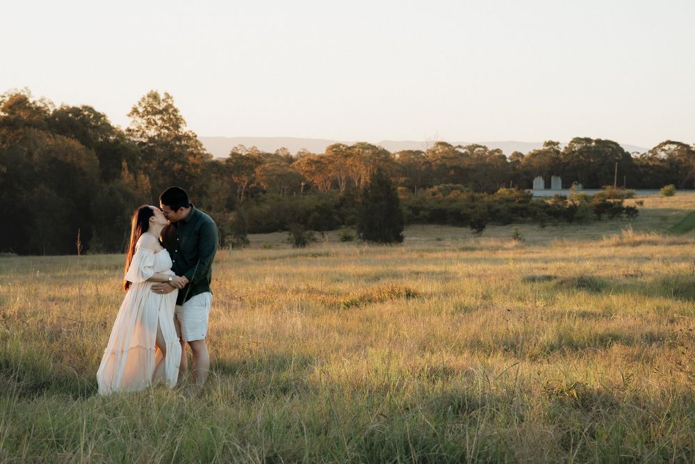 Couple embracing in a golden grassy field at sunset, the woman in a flowing white dress, with trees and a distant farmhouse behind them.
