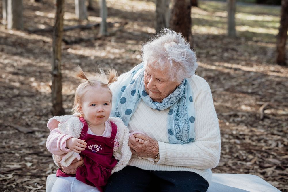 Elderly woman in a white sweater and blue scarf sits outdoors in a wooded area holding a smiling toddler in a burgundy dress in Rouse Hill.