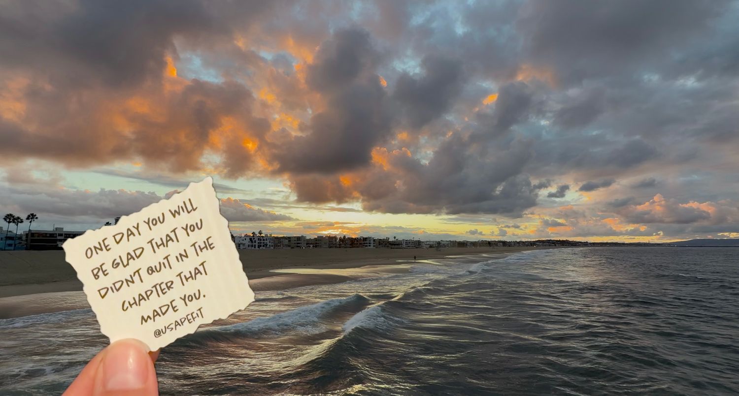 Hand holding a handwritten note about gratitude in front of a dramatic orange and gray sunset sky over ocean waves and a distant beach.