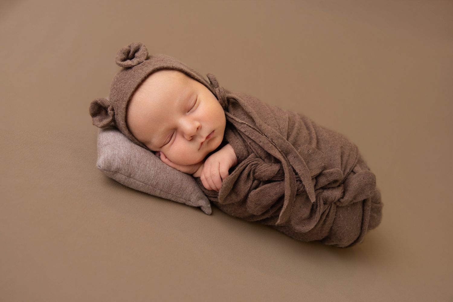 Sleeping newborn baby wrapped in a brown swaddle and bear-ear hat, resting on a small gray pillow against a soft brown backdrop.