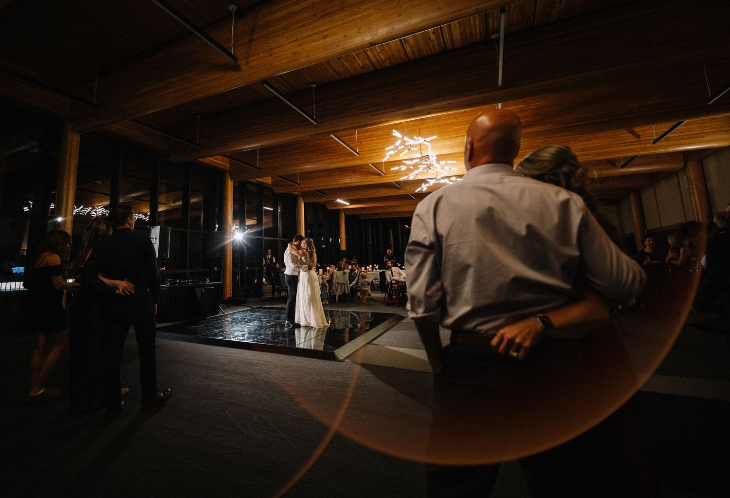 First dance under a timber ceiling framed by a glowing ring of light, guests dancing nearby, by a Calgary wedding photographer
