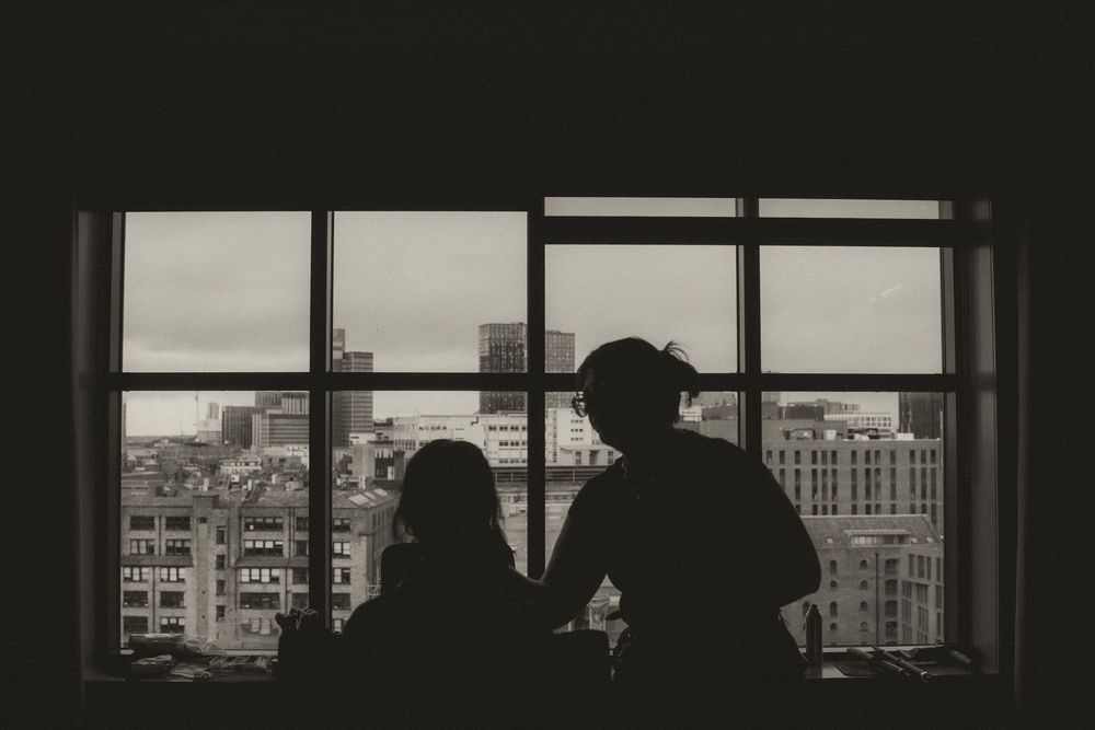 Silhouettes of two people standing before a large window overlooking a cityscape in black and white.