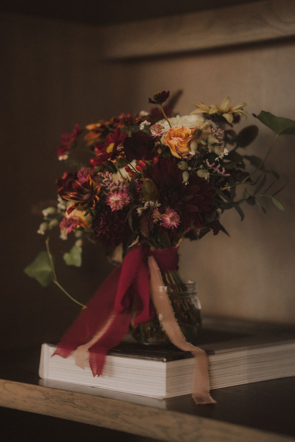A lush bouquet of burgundy, peach, and cream flowers in a mason jar with red ribbon, resting on books on a wooden shelf.