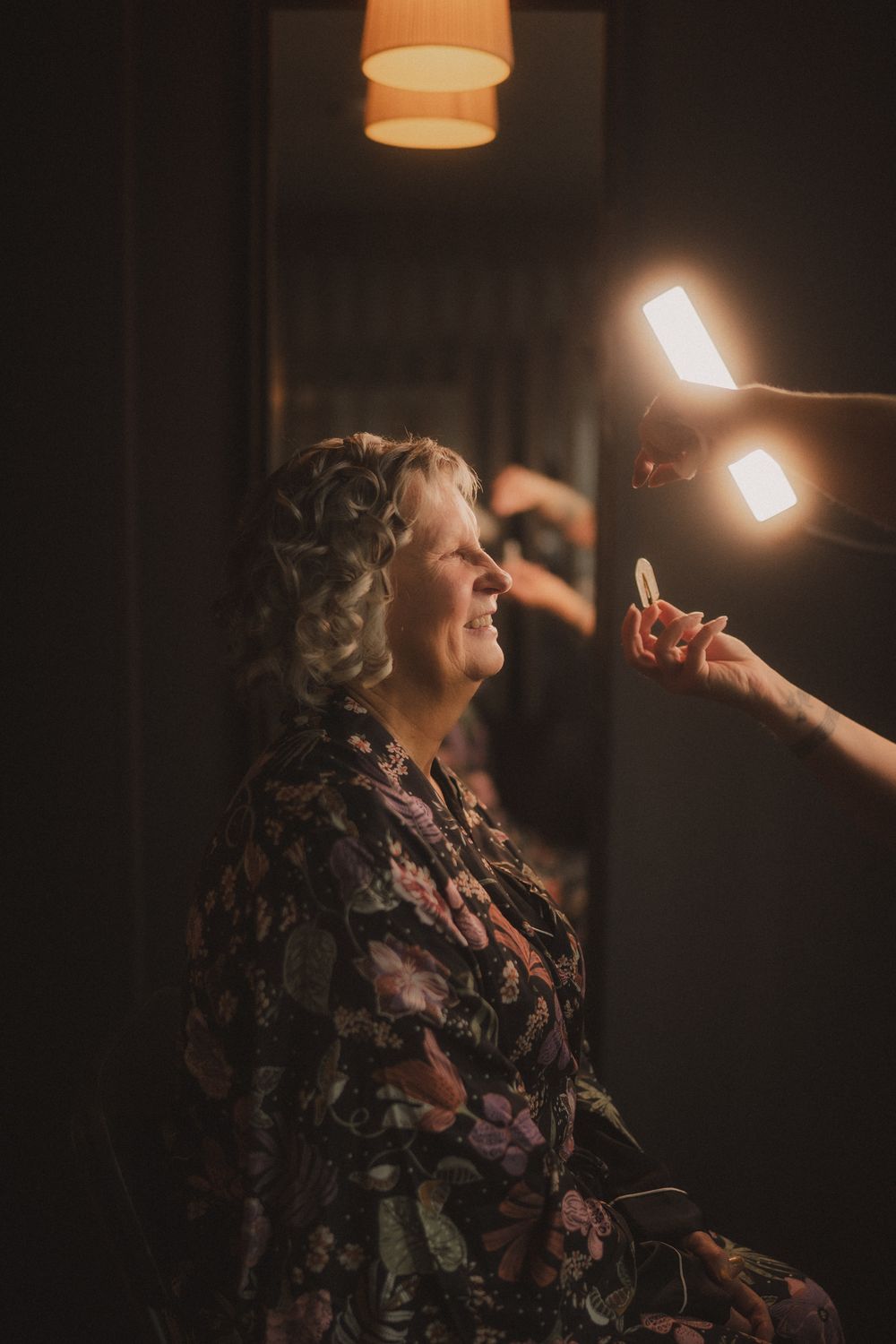 Elderly woman in floral robe getting makeup done under bright light in a dimly lit room.