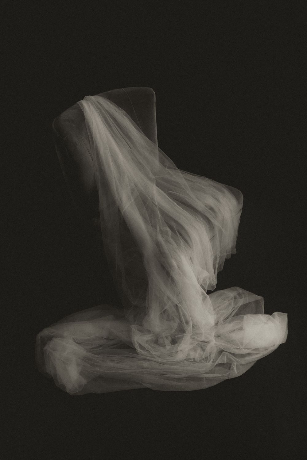 Ethereal white tulle fabric draped over a chair against a dark black background, creating a ghostly, elegant composition.