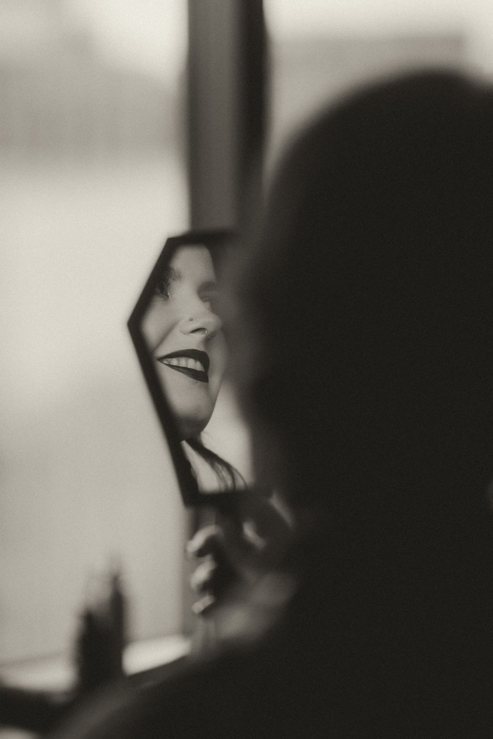 Black and white photo of a smiling woman reflected in a small mirror, holding it up to her face.