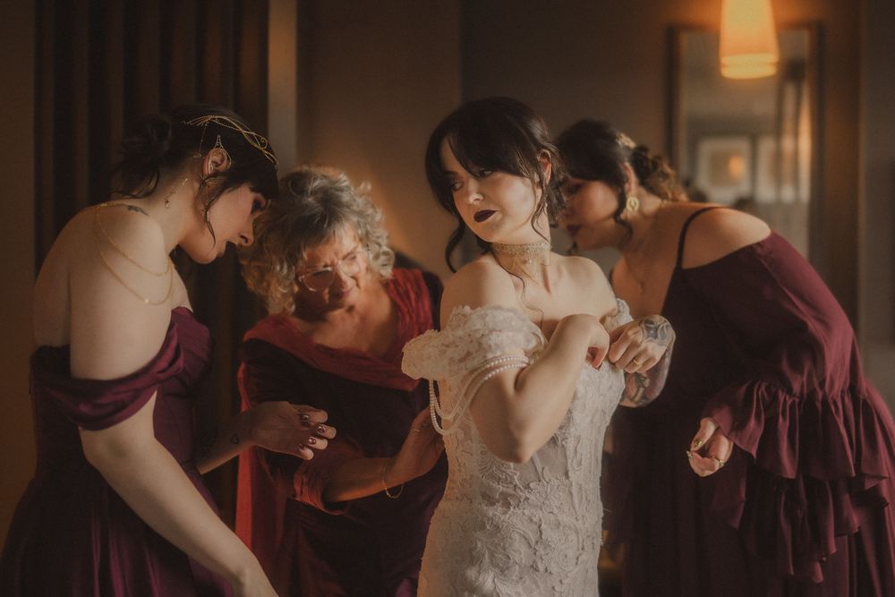Bridesmaids in burgundy dresses help a bride in a white lace gown prepare in a warmly lit room.
