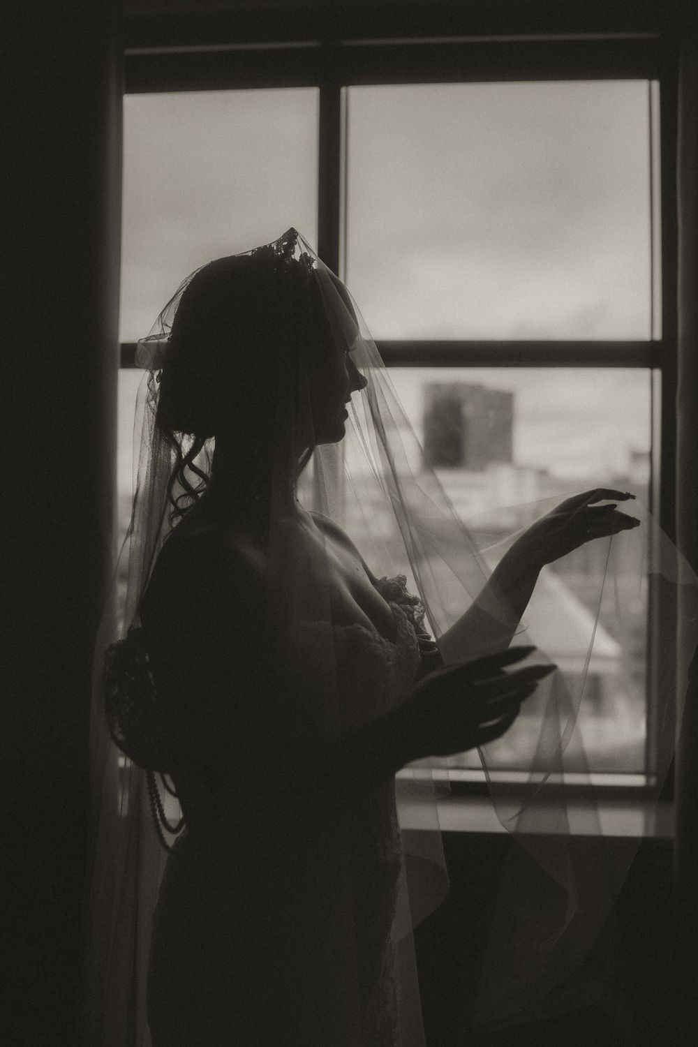 Silhouette of a bride in veil and gown standing by a window in black and white wedding photography.