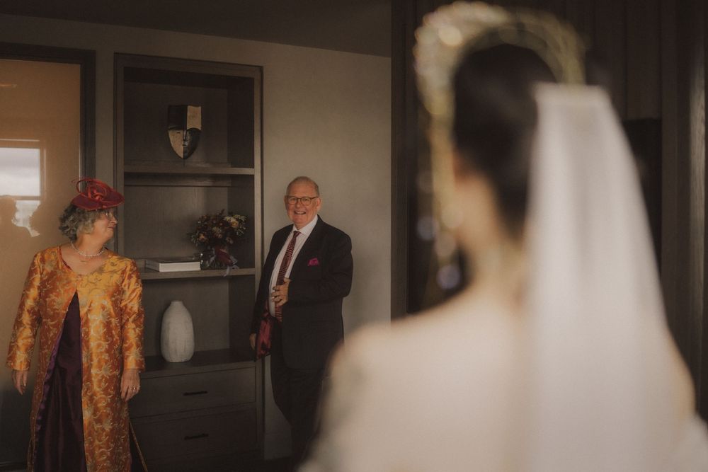 Bride in white gown and veil seen from behind, facing her parents dressed in formal attire in a warmly lit room.