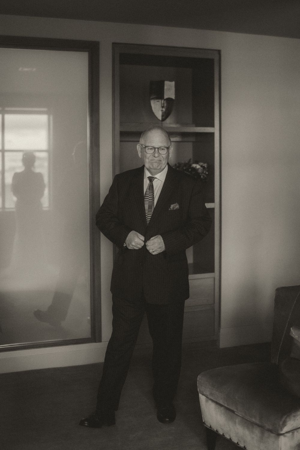 Elderly man in a dark suit adjusting his jacket, standing in a doorway in a vintage black and white photo.
