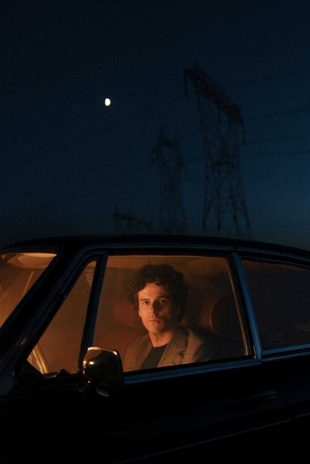 Man with curly hair sitting in a dark car at night with a full moon and power lines in the background.