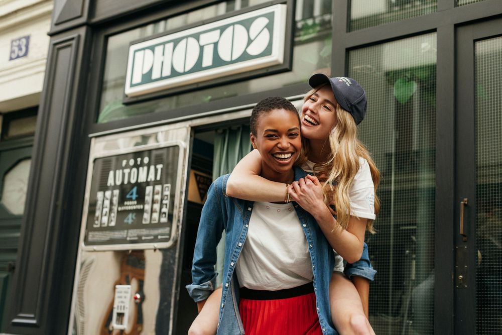 Two joyful young women laughing outside a photo booth shop, one giving the other a piggyback ride on a city street.