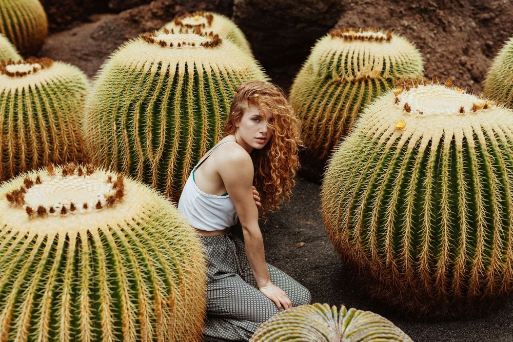 A curly-haired woman sits among large golden barrel cacti in a lush desert garden, wearing a blue top and gray pants.
