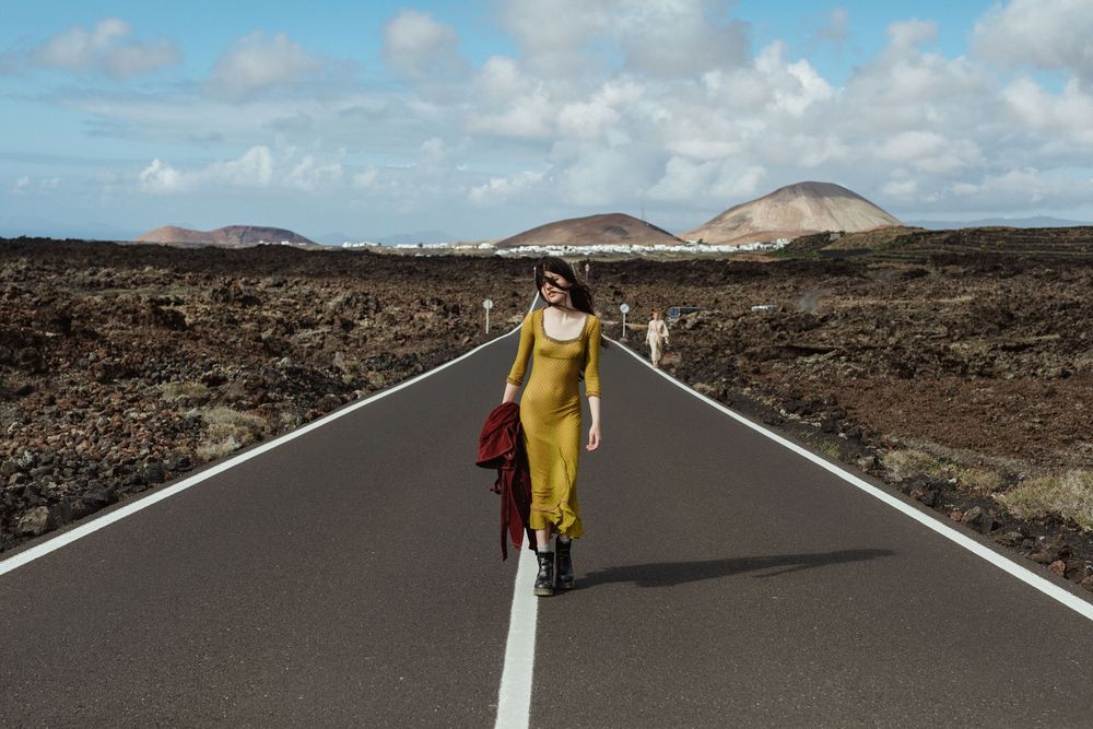 Woman in yellow dress walking on empty road through volcanic landscape with mountains under blue sky.