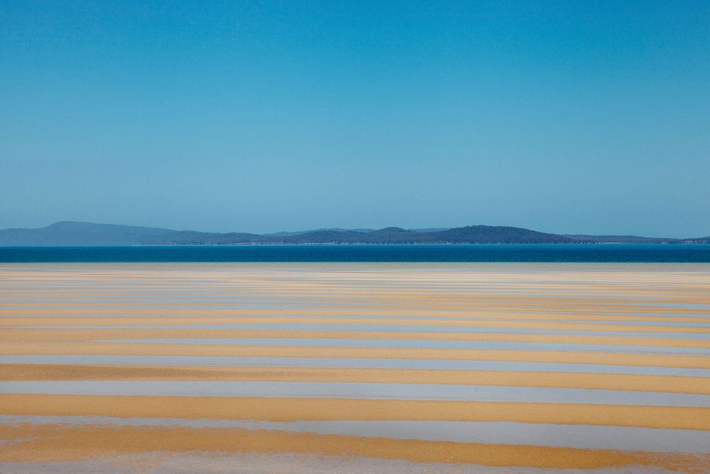 Serene low-tide beach with golden sand ripples, calm blue ocean, and distant islands under a clear sky.