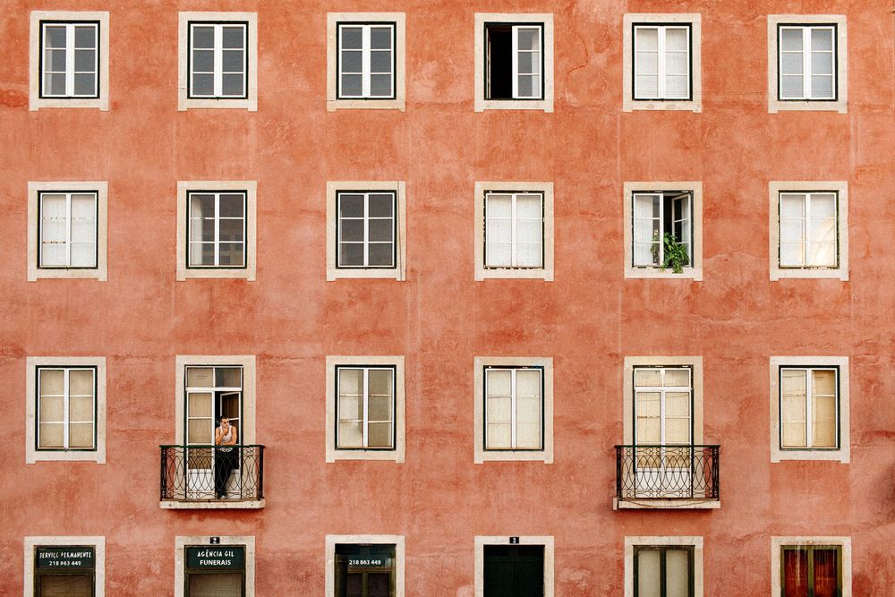 A person stands on a balcony of a terracotta-colored building with symmetrical white-framed windows in Lisbon.