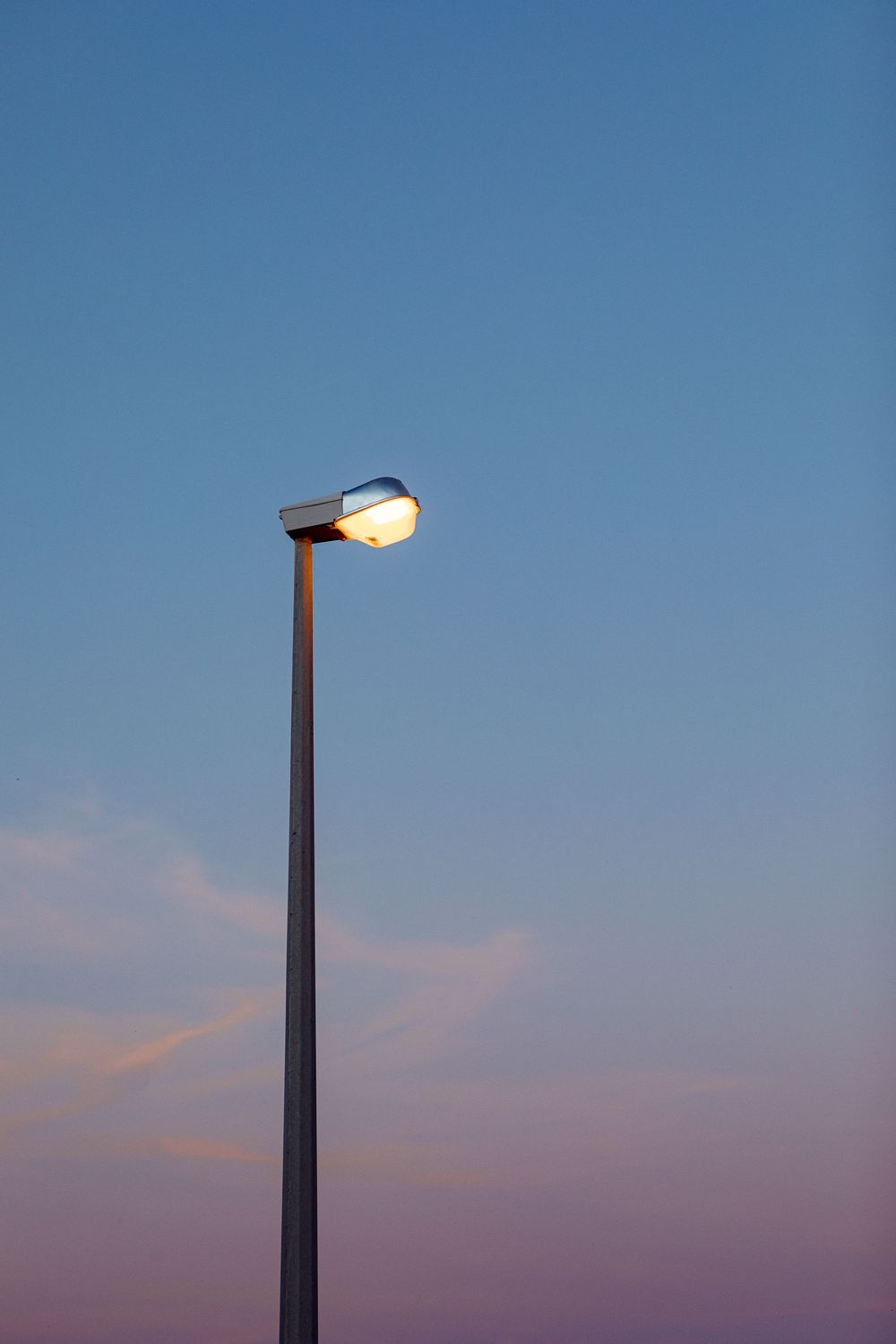 A single illuminated street lamp glowing against a serene twilight sky with soft pink and blue hues at dusk.