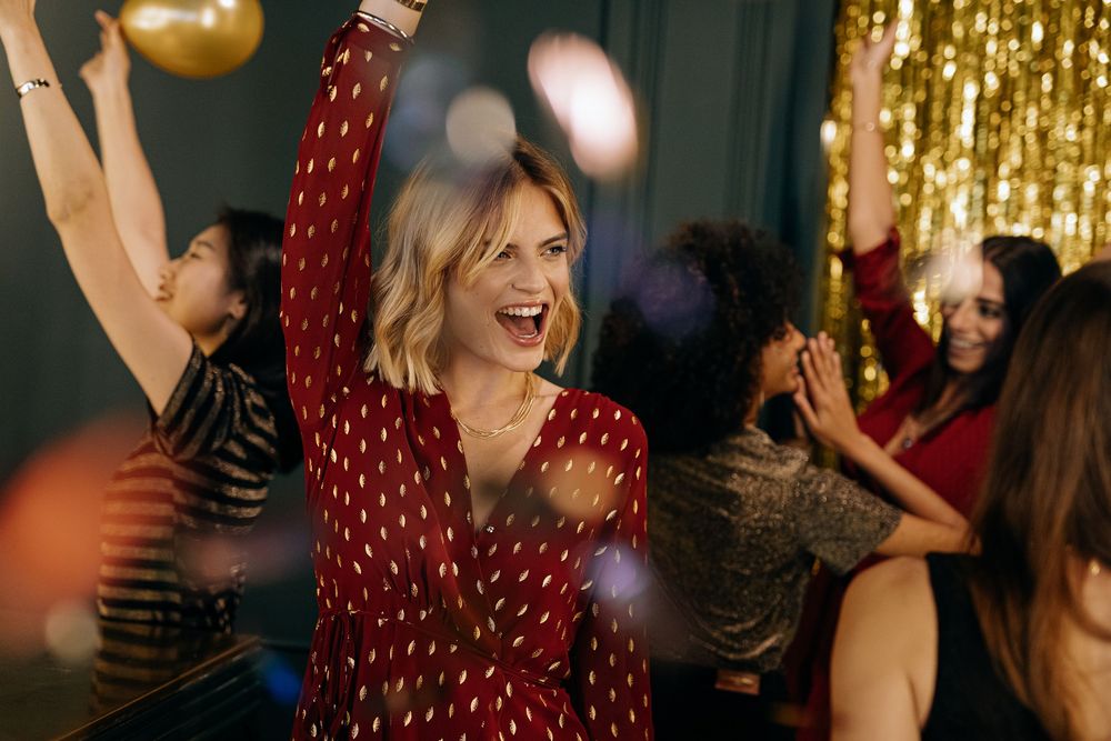 Women dancing joyfully at a party with gold decorations, colorful balloons, and festive lighting in the background.